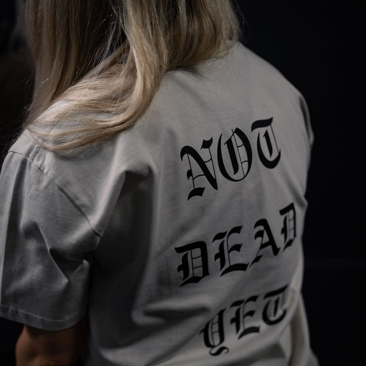 Person wearing a shirt with 'NOT DEAD YET' text on the back against a dark background