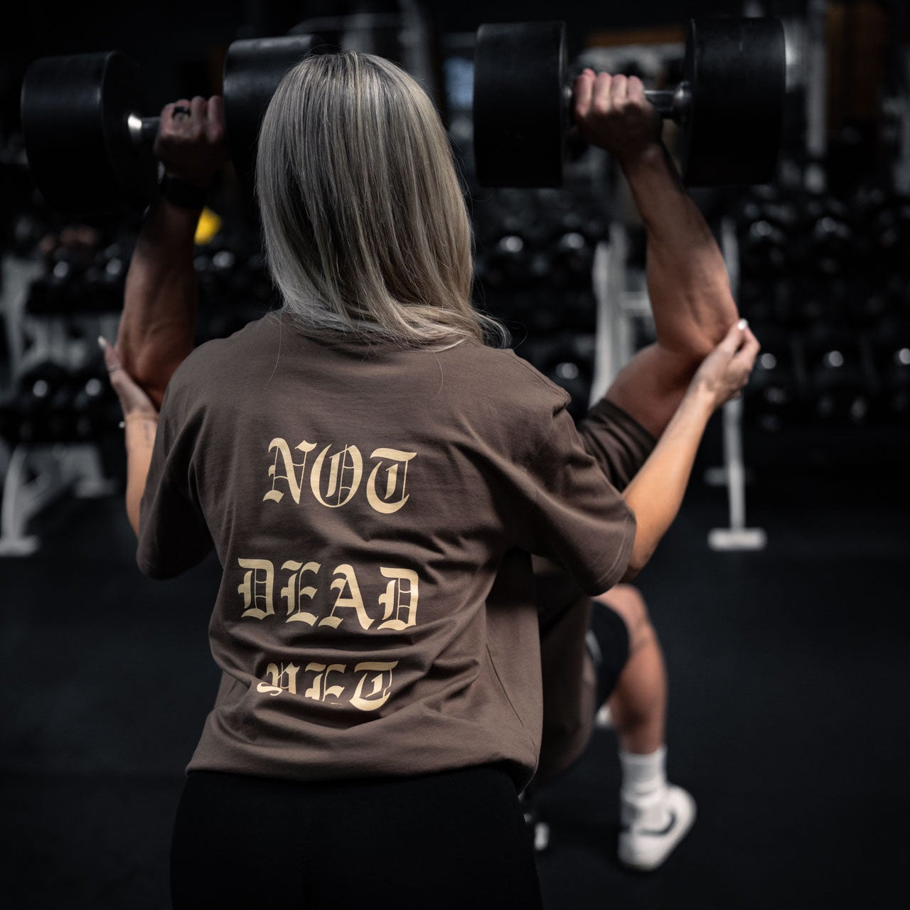 Person lifting weights in a gym wearing a shirt with 'Not Dead Yet' text.