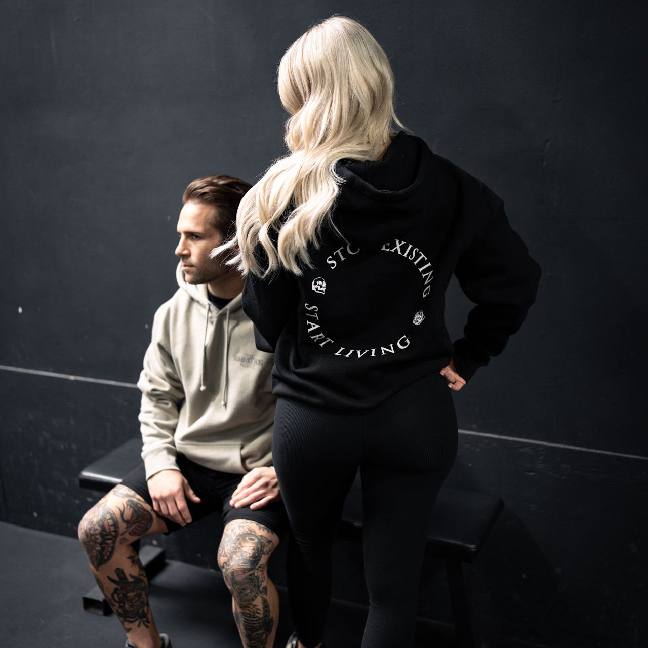 Two people sitting on a black bench against a dark background, one wearing a black hoodie with visible branding.