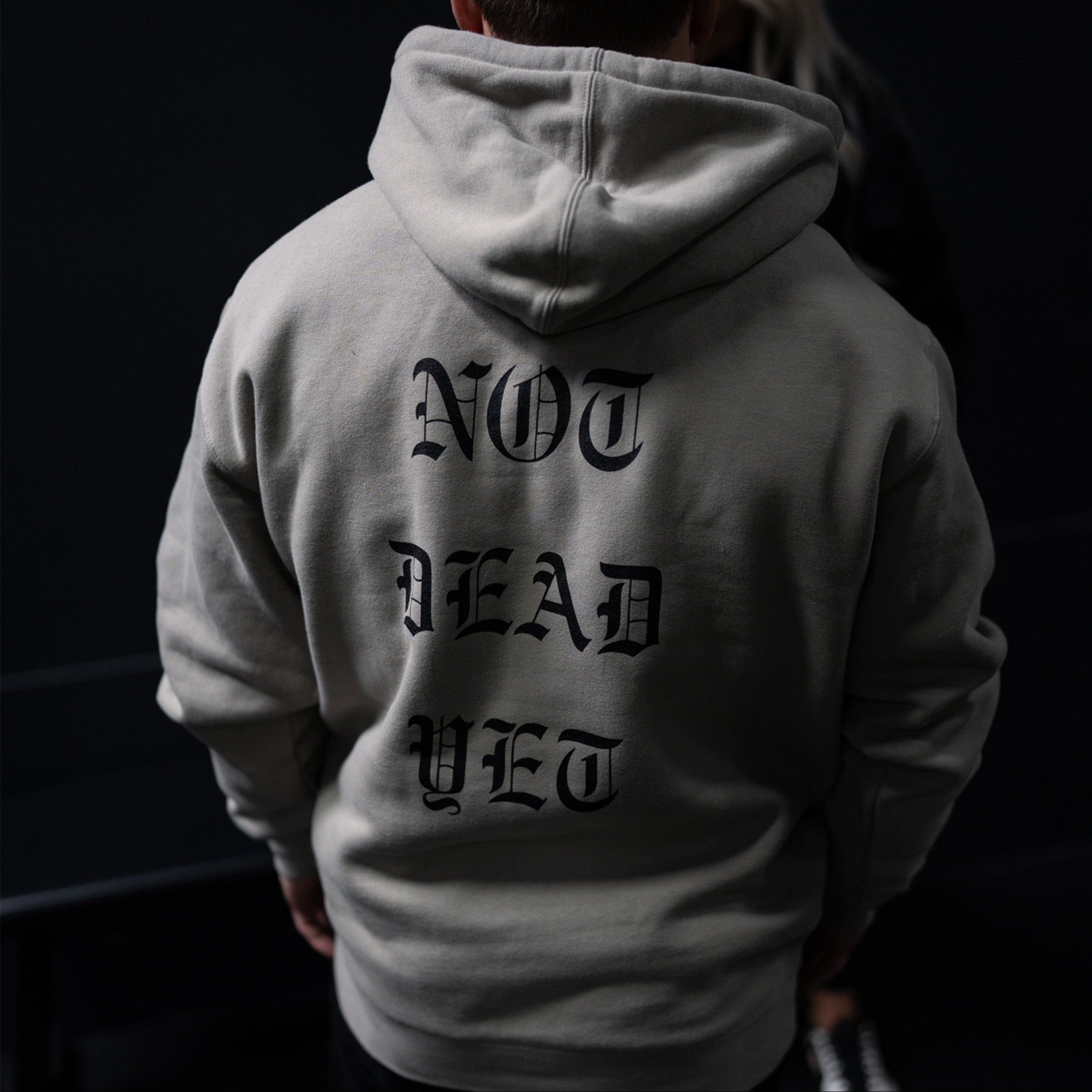 Person wearing a hoodie with 'Not Dead Yet' text on the back against a dark background