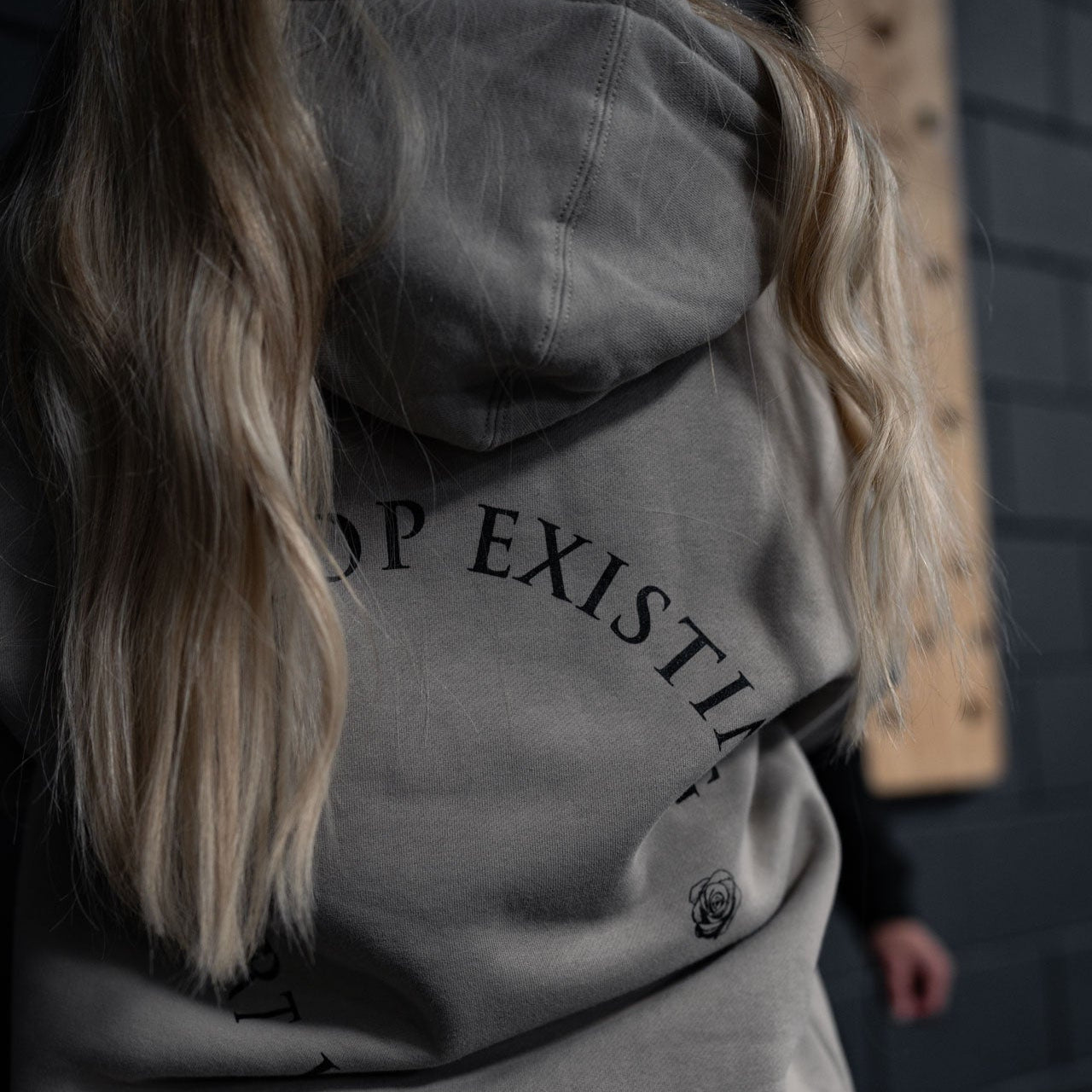 Person wearing a gray hoodie with text on the back, standing against a dark wall.