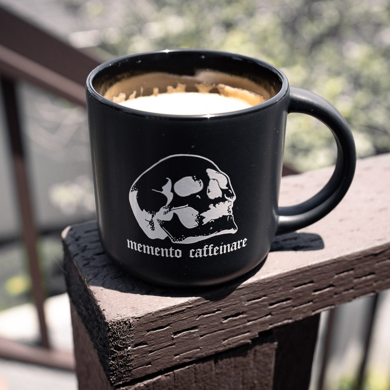 Black mug with 'memento caffeinare' design on a wooden surface outdoors