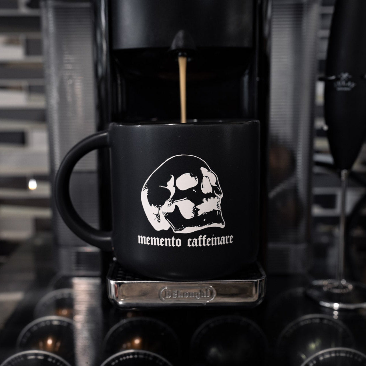 Black coffee mug with skull design and 'memento caffeinare' text being filled with coffee from a coffee machine.